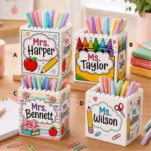 May include: Four white square desk organizers made of building blocks, each filled with colorful pens. They feature personalized labels: Mrs. Harper with an apple and pencil, Ms. Taylor with crayons, Mrs. Bennett with books, and Ms. Wilson with a pencil.