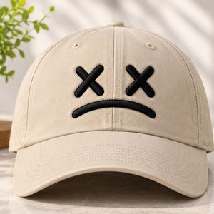 May include: A beige baseball cap featuring an embroidered black graphic on the front panel. The design consists of two black X shapes positioned above a curved black line, resembling a sad face with crossed-out eyes on the solid fabric.