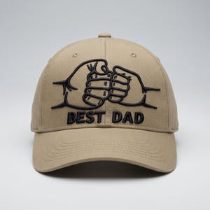 May include: A tan baseball cap featuring a black embroidered graphic of two fists performing a fist bump. Below the fist bump graphic, the text BEST DAD is embroidered in black, bold, capital letters. The cap has a curved brim and eyelets.
