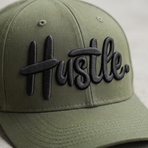 May include: An olive green baseball cap featuring the word Hustle embroidered in raised black lettering across the front panels. The cap is constructed from a durable twill fabric with matching stitching and a structured crown design.