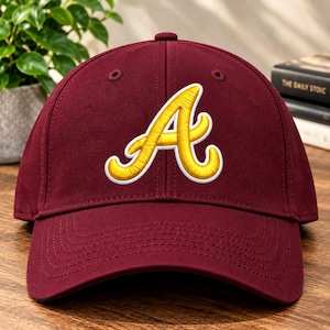 Puede incluir: Una gorra de béisbol color granate con una letra A bordada en amarillo brillante y contorno blanco. La gorra presenta una corona estructurada, una visera curva y ojales cosidos en los paneles superiores, con un diseño deportivo clásico.