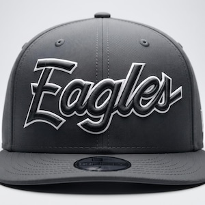 May include: A charcoal grey baseball cap featuring the word Eagles embroidered in a raised, stylized black font with a white outline across the front panels. The cap has a flat brim and a circular sticker centered on the top of the brim.