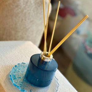 Könnte beinhalten: Eine dunkelblaue, zylindrische Glasflasche für einen Duftdiffusor mit silberfarbenem Hals und drei hellbeigen Stäbchen. Die Flasche steht auf einem durchscheinenden Untersetzer mit gewelltem Rand, der blaue Glitzerpartikel und metallische Splitter enthält.