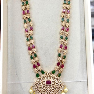 May include: A gold-toned necklace and earring set featuring teardrop-shaped green and pink stones, clear crystals, and small white pearls. The necklace has a large, intricate central pendant, while the earrings are bell-shaped jhumka style.