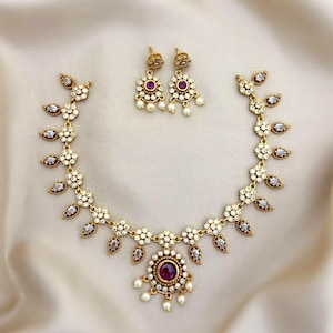 May include: A gold-toned necklace and matching earring set featuring clear white stones and a central deep red stone. The necklace has a floral-inspired link design with dangling white beads, while the earrings mirror the central pendant design.