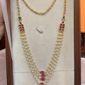 May include: A multi-strand necklace featuring small white pearls and gold beads. The design includes square green stones, clusters of teardrop-shaped red stones, and four oval-shaped red beads centred at the bottom of the cascading pearl strands.