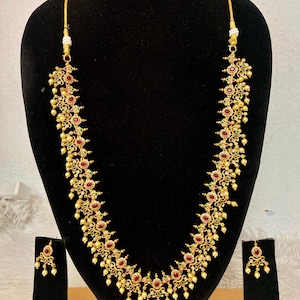 May include: A long gold-tone necklace featuring intricate metalwork, embedded with small red and green stones, and accented with dangling cream-colored beads. The set includes a matching pair of drop earrings with the same stone and bead design.