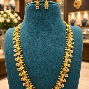 May include: A long gold necklace featuring repeating floral motifs and small deity figures, accented with tiny red and green stones. The set includes a matching pair of gold stud earrings, each displaying a central deity figure with red and green stone accents.