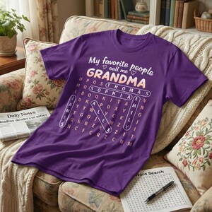May include: A purple short-sleeved t-shirt featuring the white text My favorite people call me GRANDMA above a word search puzzle grid. The grid highlights names including THOMAS, SOPHIA, EMILY, JAMES, and ALEX in white circles.