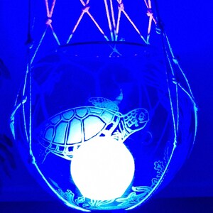 May include: A round glass bowl with an etched sea turtle design, illuminated by a bright white light source from within. The bowl is suspended by a white macrame cord hanger. The entire object is bathed in a vibrant blue light, highlighting the turtle.