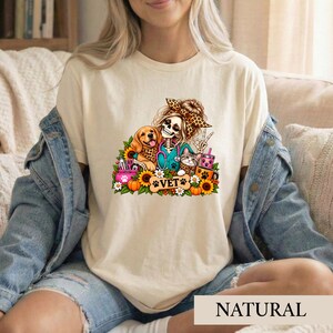 Puede incluir: Una camiseta de color crema con un estampado gráfico de un esqueleto que lleva una diadema de leopardo y una blusa de color verde azulado con un estetoscopio. El diseño incluye un golden retriever, un gato, girasoles, calabazas y el texto VET.