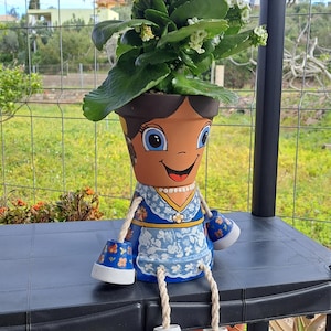 May include: A terracotta pot painted to resemble a person with a smiling face, brown hair, and a blue dress with a white floral apron. The pot features dangling arms and legs made of rope with small painted terracotta cups as hands and feet.