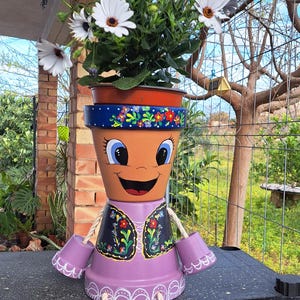 Può includere: Un vaso decorativo realizzato con vasi in terracotta impilati, dipinto come una figura dal volto sorridente, che indossa un gilet viola con motivi floreali, con arti fatti di vasi blu e corde bianche, contenente fiori di margherita bianchi.