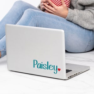 May include: A silver laptop with the name "Paisley" in teal script and a small red heart. The laptop is open and resting on a person's lap. The person is wearing blue jeans and a gray sweater.
