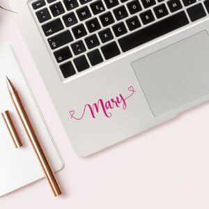 May include: A laptop with a keyboard and trackpad. A pink sticker with the name "Mary" in cursive script is on the laptop. Two small hearts are on either side of the name. A notebook and pen are in the background.