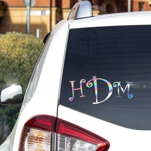 K&ouml;nnte beinhalten: Ein wei&szlig;es Auto mit einem Aufkleber an der Heckscheibe, auf dem "HDM" in einer regenbogenfarbenen, schillernden Schrift steht.