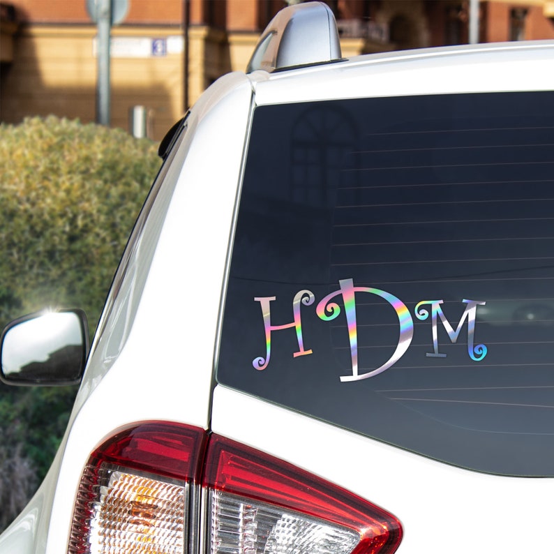 K&ouml;nnte beinhalten: Ein wei&szlig;es Auto mit einem Aufkleber an der Heckscheibe, auf dem "HDM" in einer regenbogenfarbenen, schillernden Schrift steht.