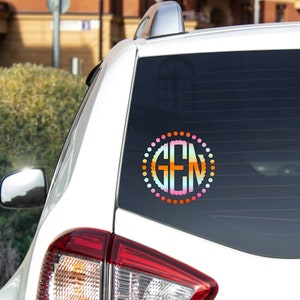 K&ouml;nnte beinhalten: Ein wei&szlig;es Auto mit einem bunten Monogramm-Aufkleber an der Heckscheibe. Der Aufkleber ist ein Kreis mit wei&szlig;em Hintergrund und einem Regenbogen-Ombre-Effekt. Die Buchstaben "GEN" befinden sich in der Mitte des Kreises in wei&szlig;er Schrift.