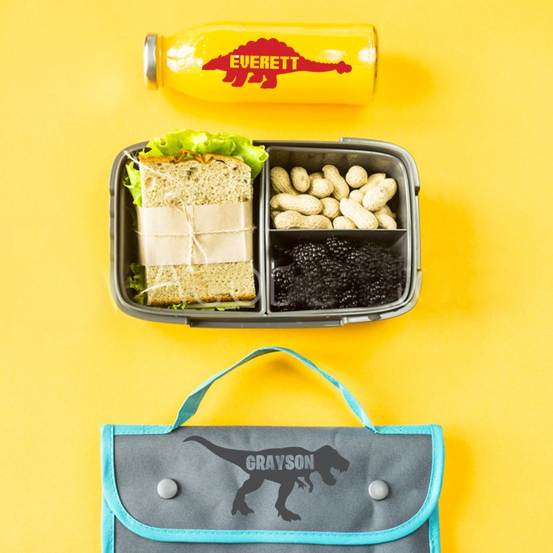 Op de afbeelding: Een grijze lunchtas met een blauwe rits en een dinosaurusafbeelding met de tekst "Grayson". Een grijze lunchbox met een sandwich, pinda's en bramen. Een gele waterfles met een rode dinosaurusafbeelding met de tekst "Everett".