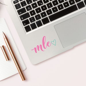 May include: A silver laptop with a pink and blue sticker that says "mle" with a heart. The laptop is on a pink surface.