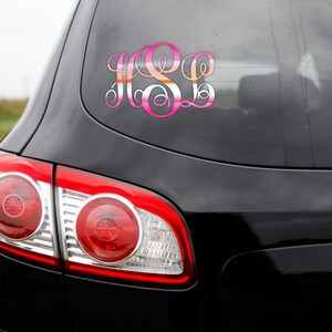K&ouml;nnte beinhalten: Ein schwarzes Auto mit einem pinkfarbenen und orangen Monogramm-Aufkleber an der Heckscheibe.