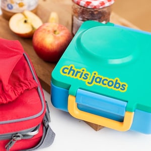 Pu&ograve; includere: Una lunch box blu e verde con una maniglia gialla. La lunch box ha un adesivo sul davanti che dice "chris jacobs".