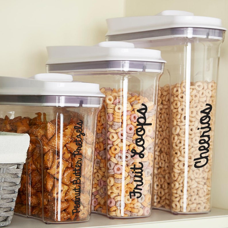 May include: Three clear, rectangular food storage containers with white lids. The containers are filled with peanut butter pretzels, colorful fruit loops, and Cheerios. Black script labels identify the contents.