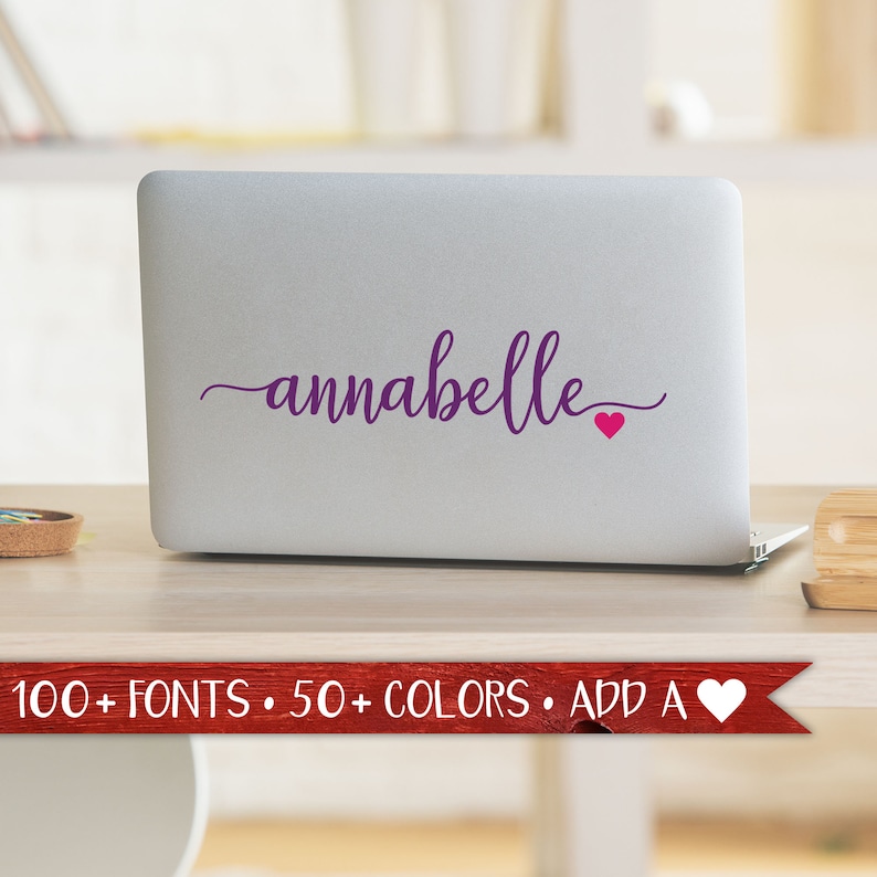 May include: A laptop with the name "annabelle" in a cursive font, in a deep purple color, with a small pink heart. The laptop is on a wooden desk. A banner at the bottom reads "100+ FONTS &bull; 50+ COLORS &bull; ADD A".