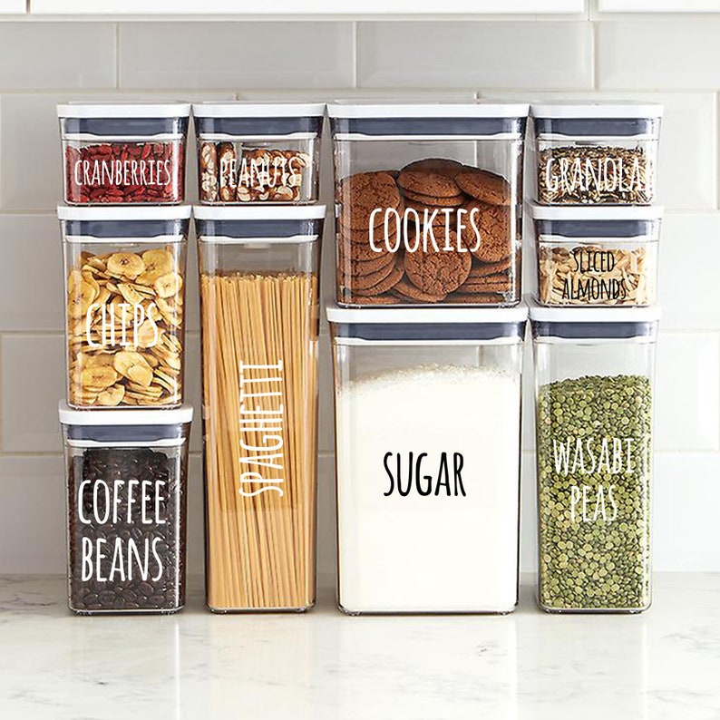 May include: A collection of clear, rectangular food storage containers with white lids. The containers hold various items like cranberries, peanuts, cookies, granola, sliced almonds, banana chips, spaghetti, coffee beans, sugar, and wasabi peas. Text labels are on each container.