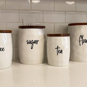 May include: Four white ceramic kitchen canisters with wooden lids, labeled "coffee", "sugar", "tea", and "flour" in black script. The canisters have a textured surface and are arranged on a white countertop.