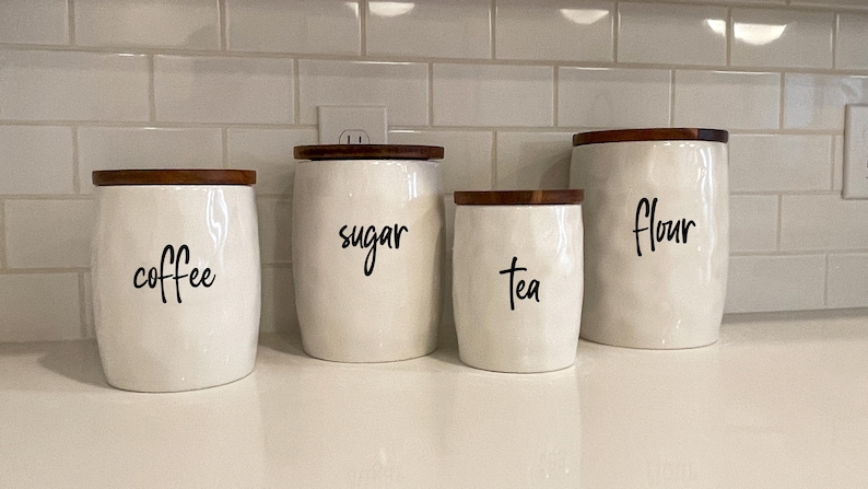 May include: Four white ceramic kitchen canisters with wooden lids, labeled "coffee", "sugar", "tea", and "flour" in black script. The canisters have a textured surface and are arranged on a white countertop.