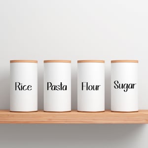 May include: Four white cylindrical kitchen canisters with wooden lids labeled "Rice", "Pasta", "Flour", and "Sugar" in black script. The canisters are arranged on a light brown wooden shelf against a white background.