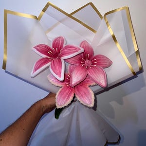 Puede incluir: Un ramo de tres flores con forma de lirio de color rosa hechas de limpiapipas difusos. Las flores presentan pétalos con bordes blancos y estambres blancos finos. El ramo está envuelto en papel blanco translúcido con bordes dorados.