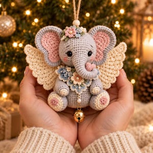 May include: A crocheted gray elephant ornament with large pink-lined ears, cream-colored wings, and a floral collar. The elephant features a small pink heart on its trunk, a flower crown on its head, and a gold jingle bell hanging from its chest.