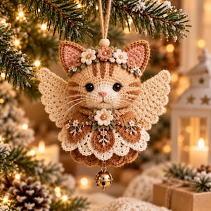 May include: A crocheted ornament shaped like a tan tabby cat with angel wings, wearing a floral crown and a decorative collar with a central flower. The cat has black eyes, a pink nose, and a small gold-colored jingle bell hanging from the bottom.