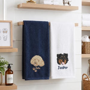 May include: A navy blue towel featuring an embroidered golden doodle and the name Mala with paw prints, and a white towel featuring an embroidered black and tan dog face and the name Jasper, both hanging on a wooden towel bar in a bathroom.
