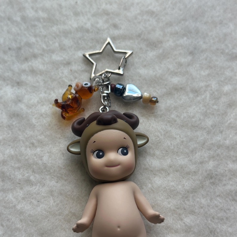 May include: A small, nude figurine with a brown ram's head covering, blue eyes, and a smiling expression. It is attached to a silver keychain with a star charm, a small glass animal, and decorative beads. The figurine is approximately 3 inches tall.