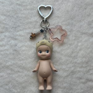 May include: A small, nude figurine with a pig hat, attached to a silver keychain with a heart-shaped clasp. The keychain also features a star charm and a pink star-shaped charm. The figurine has blue eyes and is standing upright.