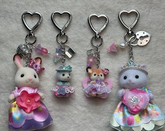 May include: Four adorable animal keychains with pastel pink, blue, and white dresses. Each keychain features a different animal: a rabbit, a cat, a deer, and a bear. The keychains are decorated with flowers, bows, and other embellishments.