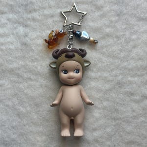 May include: A nude-colored figurine wearing a brown bull costume, with a silver star-shaped keychain. The keychain also has a small orange animal charm and decorative beads. The figurine has blue eyes and a smiling expression.
