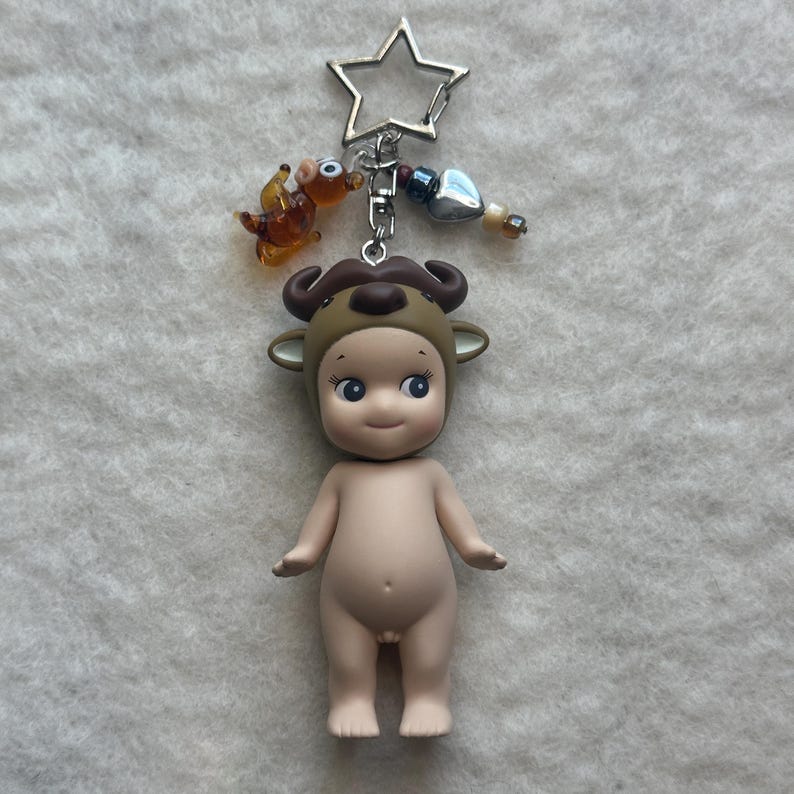 May include: A nude-colored figurine wearing a brown bull costume, with a silver star-shaped keychain. The keychain also has a small orange animal charm and decorative beads. The figurine has blue eyes and a smiling expression.