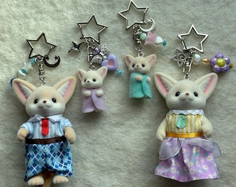 May include: Three adorable fox keychain charms with different outfits and accessories. The fox on the left is wearing a blue and white striped shirt with a red tie and blue plaid pants. The fox in the middle is wearing a lavender sweater. The fox on the right is wearing a yellow and white striped shirt with a green bow and a lavender skirt with white flowers.