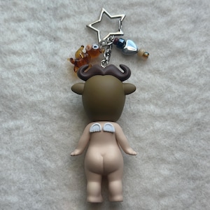 May include: A novelty keychain featuring a figurine with a bull-like head, a mustache, and small white wings. The keychain includes a star-shaped charm, a small glass animal, and decorative beads. The figurine is nude with a light skin tone.