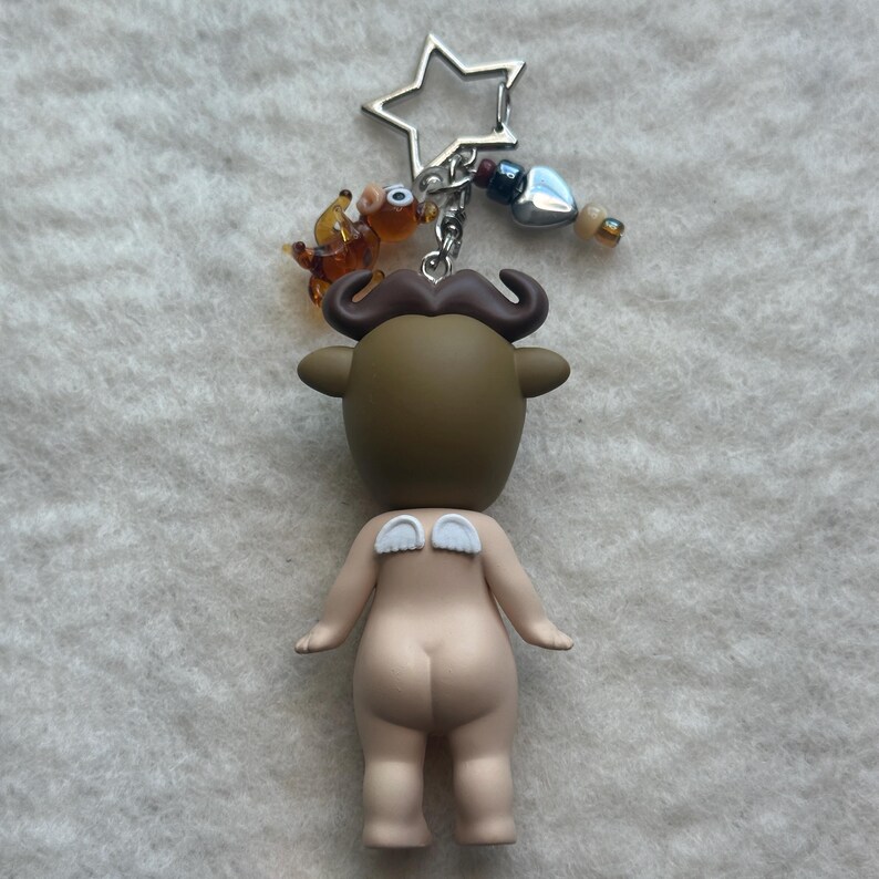 May include: A novelty keychain featuring a figurine with a bull-like head, a mustache, and small white wings. The keychain includes a star-shaped charm, a small glass animal, and decorative beads. The figurine is nude with a light skin tone.