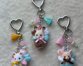 May include: Three plush animal keychains with heart-shaped key rings. Each keychain features a different animal: a brown rabbit, a white mouse, and a tan fox. Each animal is wearing a blue and pink scarf and a pink bow. The keychains are decorated with beads and tassels.