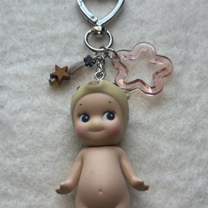 May include: A small, nude doll keychain with a pig hat, attached to a silver-colored clasp. The keychain includes a pink star-shaped charm and a wooden star charm. The doll has large blue eyes and rosy cheeks.