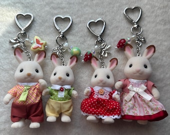 May include: Four white rabbit figurines with red, pink, green, and yellow clothing. Each figurine is attached to a silver heart-shaped keychain with charms.