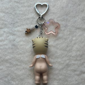 May include: A keychain featuring a small figurine with a tiger head and angel wings. The figurine is attached to a silver heart-shaped clasp with a pink star and a gold star charm. The figurine is tan with black stripes.