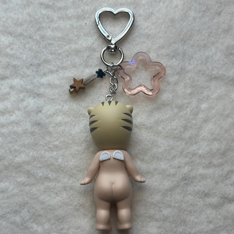 May include: A keychain featuring a small figurine with a tiger head and angel wings. The figurine is attached to a silver heart-shaped clasp with a pink star and a gold star charm. The figurine is tan with black stripes.