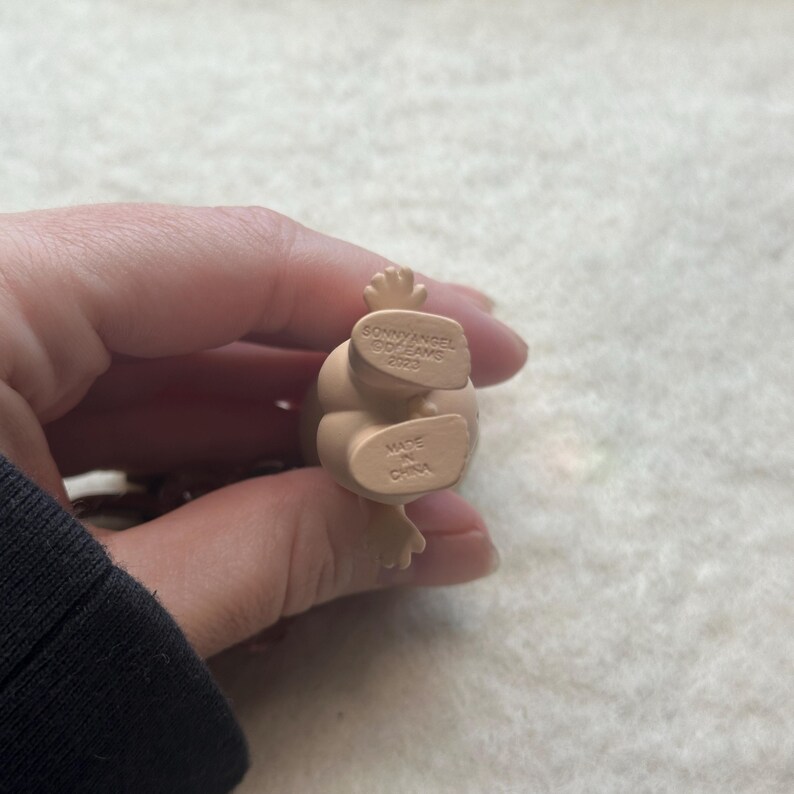 May include: A small, beige Sonny Angel figurine. The bottom of the figurine has the text "SONNY ANGEL DREAMS 2023" and "MADE IN CHINA". The figurine is held in a person's hand.